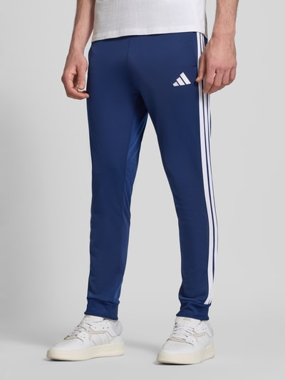 Mężczyzna nosi 'Trainingsanzug mit elastischen Abschlüssen' od adidas Sportswear w kolorze niebieskim z białymi detalami, detaliczny widok nóg, krój prosty, elastyczne wykończenia, logo, spodnie dresowe z elastycznym pasem, wpuszczane kieszenie, szerokość w rozmiarze M: 36 cm, długość wewnętrzna nogawki: 75 cm.