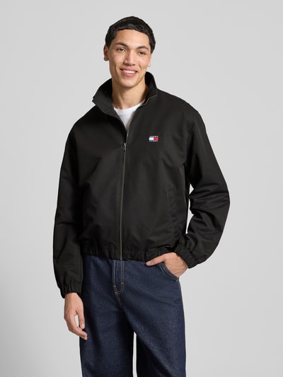 Männermodel trägt Regular Fit Blouson van Tommy Jeans in effen zwart met opstaandekraag, label patch, lange mouwen, elastische boorden, zijzakken, geheel vooraanzicht.