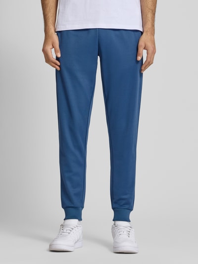 Mężczyzna nosi spodnie dresowe Sweatpants mit elastischem Bund Modell 'BERTONI' od Ellesse, mieszanka bawełny, krój regular fit, detal z logo, długość wewnętrzna nogawki w rozmiarze M: 76 cm, kolor niebieski, gładka faktura, całkowity widok z przodu.