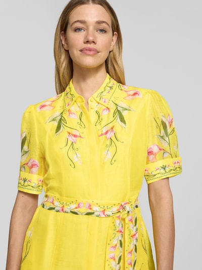 Modelka kobieca nosi Minikleid aus reiner Viskose mit floralem Allover-Print von Marc Cain, żółty kolor, detaliczne wzory kwiatowe, taliowany krój, zdejmowany pasek materiałowy, krótkie rękawy, kołnierzyk wykładany, pełna listwa guzikowa, Szerokość w rozmiarze 36: 41 cm, długość pleców 93 cm, Szerokość w rozmiarze 36: 41 cm, długość rękawa 29 cm; zbliżenie na górną część ciała.