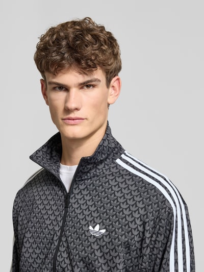 Männermodel trägt Sweatjacke mit Stehkragen von adidas Originals mit Label-Stitching, Reißverschlusstaschen, gerippten Abschlüssen, Nahaufnahme Oberkörper. Lockerer-Schnitt, Rückenlänge 72 cm, Ärmellänge 65 cm, dunkle Grundfarbe mit Muster.