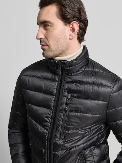 Męski model ma na sobie Regular Fit Steppjacke mit Stehkragen od Tom Tailor, czarna, jednokolorowa, krój regular fit, kołnierz stójka, zamek błyskawiczny, aplikacja, detal z logo, pasek w kontrastowym kolorze, długie rękawy z elastycznym mankietem, dół z elastycznym wykończeniem, funkcja ocieplająca, wodoodporna, boczne kieszenie na zamek, kieszeń na piersi; zbliżenie na górną część ciała z przodu.