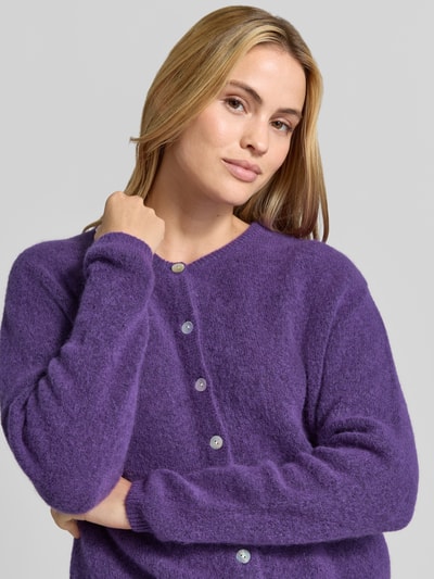 Frauenmodel trägt Relaxed Fit Strickjacke aus Woll-Mix mit Leinen-Anteil von American Vintage in Lila mit gerippten Bündchen, Nahaufnahme Oberkörper.