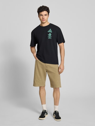 Mężczyznamodel nosi męski t-shirt z nadrukiem i okrągłym dekoltem od Edwin, czarna bawełna, krój relaxed fit, długość tyłu 72 cm, widok całej sylwetki z przodu.