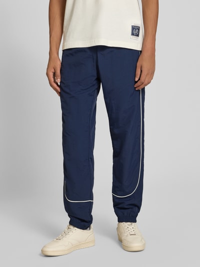 Männermodel trägt Relaxed Fit Tracksuit mit Logo-Patch von Champion in Dunkelblau mit weißen Details, strukturierte Textur, Nahaufnahme Unterkörper. Kontext: Herren Tracksuit, Relaxed Fit, elastische Abschlüsse, Innenbeinlänge 72 cm.