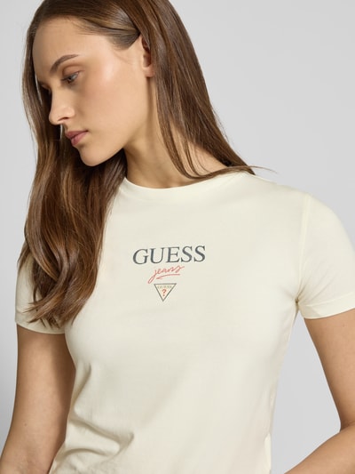 Frauenmodel trägt T-Shirt mit Logo und Rundhalsausschnitt von Guess Jeans im Slim Fit, Farbton Creme, Baumwoll-Elasthan-Mix, Nahaufnahme Oberkörper.