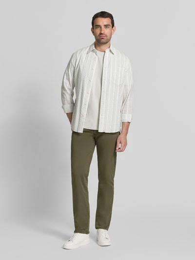 Männermodel trägt olivgrüne Stoffhose aus Baumwoll-Mix mit Viskose-Anteil Modell 'Cadiz' von Brax, kombiniert mit heller Kleidung, Ganzkörperansicht von vorne. Herren Stoffhose von Brax.