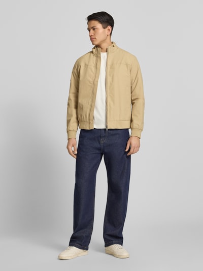 Männermodel trägt Jacke mit Stehkragen und Eingrifftaschen von Superdry, krój regular fit, zamek błyskawiczny, wpuszczane kieszenie, długość tyłu w rozmiarze M: 70 cm, szerokość ramion w rozmiarze M: 44 cm, szerokość klatki piersiowej w rozmiarze M: 41 cm, długość rękawów w rozmiarze M: 68 cm, beżowa, gładka faktura, ciemnoniebieskie spodnie, jasne buty, jasnoszare tło, Ganzkörperansicht von vorne