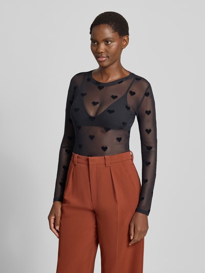 Frauenmodel trägt transparentes schwarzes Slim Fit Langarmshirt mit Herzmuster von VERO MODA, kombiniert mit rostbrauner Hose, Nahaufnahme Oberkörper.