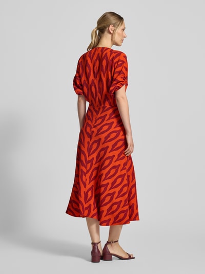 Männermodel trägt A-Linien Freizeitkleid mit V-Ausschnitt Modell 'KOMFORTABLE' von Essentiel Antwerp, czerwono-pomarańczowy wzór, tekstura gładka, Ganzkörperansicht Rückansicht, sukienki casualowe.