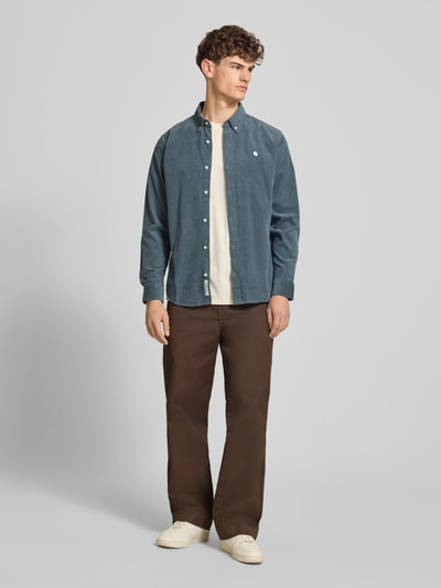 Männermodel trägt braune Hose mit Gesäßtaschen Modell 'Denison' von Carhartt Work In Progress, Material-Mix, Relaxed-Fit, Ganzkörperansicht von vorne.