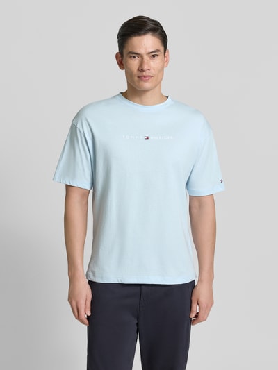 Męski model nosi Regular Fit T-Shirt mit Label-Schriftzug aus reiner Baumwolle von Tommy Hilfiger, jasnoniebieski, gładka faktura, krój regular fit, okrągły dekolt, krótki rękaw, przód nadruk z logo, bawełna, Ganzkörperansicht von vorne.