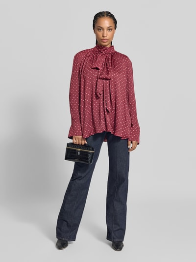 Vrouwenmodel draagt Regular Fit Bluse aus Satin van ROTATE in bordeaux met witte stippen, opstaande kraag, sierstrik, blouse met lange mouwen, ruglengte maat 36: 70 cm, mouwlengte maat 36: 63 cm, effen donkere broek, volledige lichaamsaanzicht van voren.