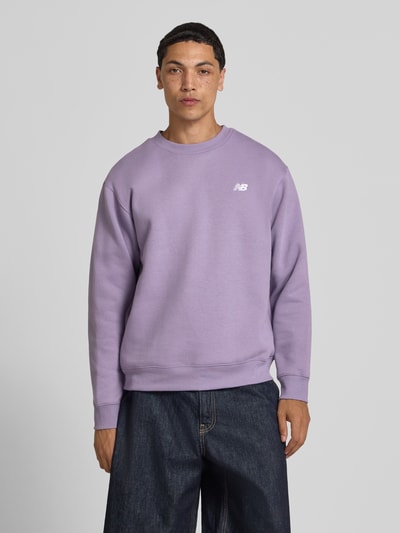 Männermodel trägt Sweatshirt mit Rundhalsausschnitt und Label-Stitching von New Balance in Lila, Baumwoll-Mix, Relaxed-Fit, Ganzkörperansicht von vorne. Rückenlänge bei Größe M 67 cm, Ärmellänge 63 cm.