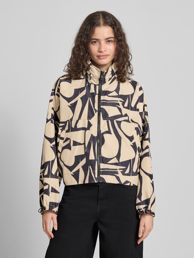 Modelka nosi Oversized Hemdjacke mit Allover-Muster Modell 'BETTY BATSLEEVE' od ONLY, beżowo-czarna z graficznym wzorem, stójka z chowaną kapturem, rękawy nietoperzowe, elastyczne końce rękawów, krótkie, wpuszczane kieszenie, zamek błyskawiczny, efekt: wzór na całej powierzchni, krój oversized, widok pełnej sylwetki od przodu.