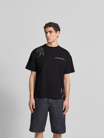 Männermodel trägt Relaxed Fit T-Shirt aus Baumwoll-Mix von Don´t Waste Culture in Schwarz mit Motiv-Print, Rundhalsausschnitt, Label-Print, Rückenlänge bei Größe M 70 cm, Ganzkörperansicht von vorne