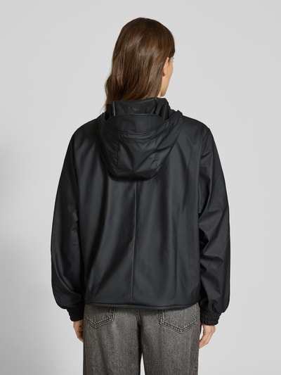 Frauenmodel trägt Jacke mit Abdeckleiste von Rains in Schwarz, gerade Passform, Kapuze mit Tunnelzug, elastische Ärmelabschlüsse, Ganzkörperansicht Rückansicht. Rückenlänge 58 cm, Schulterbreite 43 cm, Brustweite 55 cm, Ärmellänge 63 cm bei Größe S.