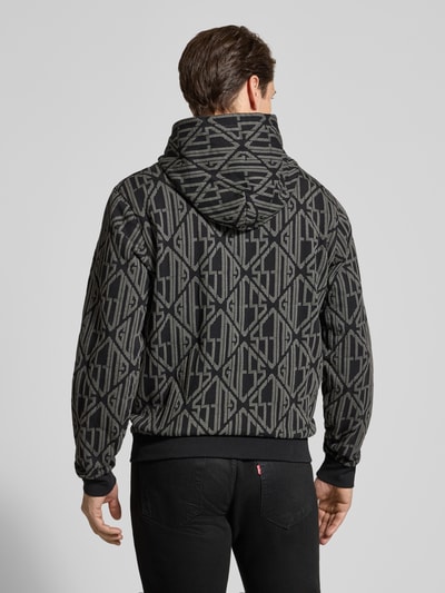 Männermodel trägt Hoodie im Allover-Look mit Känguru-Tasche von Guess, Reine Baumwolle, Regular-Fit, Rückenlänge 67 cm, Ärmellänge 67 cm, dunkle Töne, grafische Muster, Ganzkörperansicht Rückansicht.
