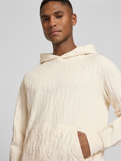 Männermodel trägt Hoodie mit Känguru-Tasche und Kapuze von Guess aus reiner Baumwolle, Loose-Fit, cremefarben, Nahaufnahme Oberkörper.