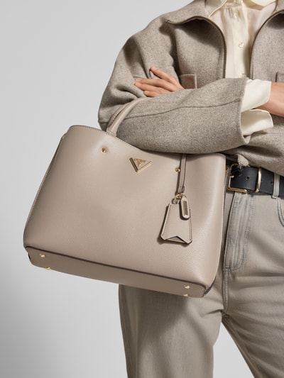 Frauenmodel trägt Handtasche mit Label-Applikation Modell 'ECO MERIDIAN II' von Guess in Beige, strukturiertes Kunstleder, Hauptfach mit Reißverschluss, Tragehenkel und Schulterriemen, Größe 33x23x11 cm, Schulterriemen 106 cm, Ganzkörperansicht von vorne.