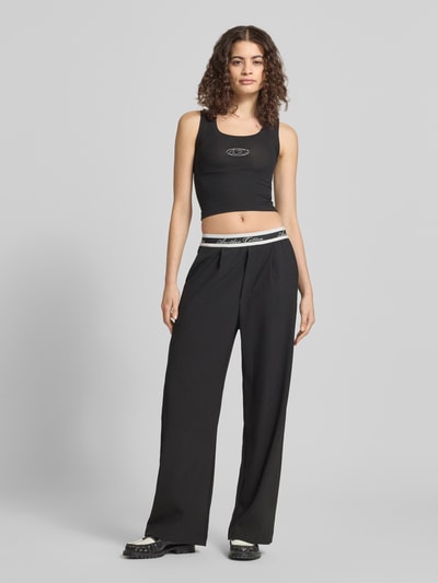 Kobiece modelka nosi spodnie z szerokimi nogawkami 'Wide Leg Hose mit elastischem Logo-Bund' od ANOTHER COTTON LAB, mieszanka materiałów z dodatkiem streczu, wpuszczane kieszenie, szerokość w rozmiarze S: 36 cm, długość wewnętrzna nogawki w rozmiarze S: 75 cm, czarny kolor, gładka faktura, elastyczny pasek z logo, widok całej sylwetki z przodu.