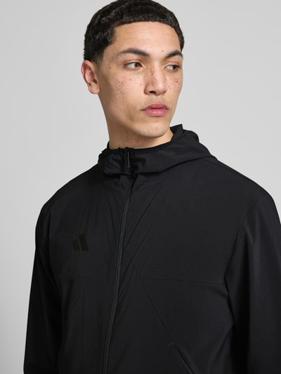 Mężczyznamodel nosi Slim Fit Funktionsjacke aus schnelltrocknendem Material von adidas Sportswear, efekt: jednolity kolor, krój slim fit, kształt kołnierza: kaptur, zapięcie: zamek błyskawiczny, aplikacja: nadruk z logo, rękawy: długi rękaw, funkcja: szybkoschnący, z regulacją wilgoci, boczna kieszeń zapinana na zamek błyskawiczny, materiał gładki, czarny kolor, nahaufnahme górnej części ciała