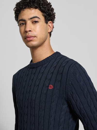 Männermodel trägt Slim Fit Pullover in Strick-Optik von Superdry aus reiner Baumwolle, Logo-Stitching, Rundhalsausschnitt, Nahaufnahme Oberkörper.