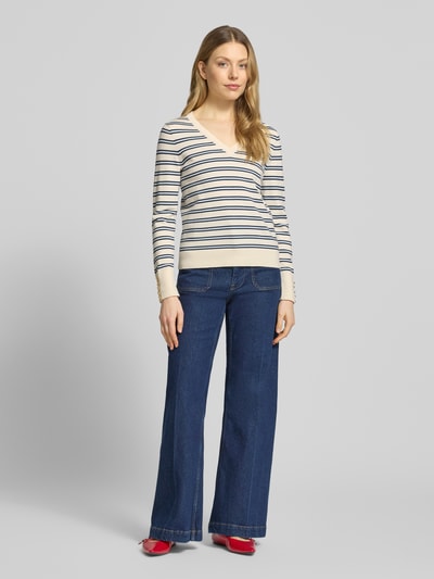 Vrouwenmodel draagt Regular Fit gebreide pullover uit puur katoen van Tommy Hilfiger met gestreepte look, gestikt logo, V-hals, lange mouw, sierknopen, geribde boorden; all-over motief, zachte textuur, kleur crème-blauw, vooraanzicht full-body.
