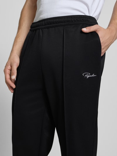 Mężczyzna nosi Trackpants mit Längsnähten Modell 'Motus' od Pegador, czarny kolor, materiał z bawełną, krój relaxed fit, elastyczny pas, długość nogawki 75 cm w rozmiarze M, zbliżenie na górną część nogawki z przodu.