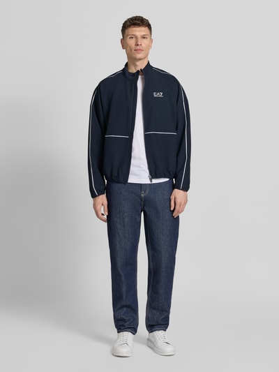 Männermodel trägt Bomberjacke mit Stehkragen und Logo-Print von EA7 Emporio Armani in Dunkelblau, gerader Schnitt, elastische Abschlüsse, Ganzkörperansicht von vorne.