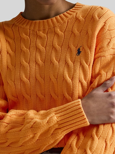 Vrouwenmodel draagt Regular Fit Strickpullover aus reiner Baumwolle van Polo Ralph Lauren in effen oranje met kabelpatroon, logo-stiksel, ronde hals en geribde boorden, gebreide textuur, materiaal katoen. Nahaufnahme bovenlichaam.