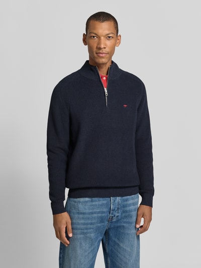 Męski model nosi Regular Fit Strickpullover aus reiner Baumwolle mit Troyer-Kragen od Fynch-Hatton, ciemnoniebieska dzianina, długi rękaw, logo, żebrowane wykończenia, widoczna struktura dzianiny, krój regular fit, widok całej sylwetki z przodu.