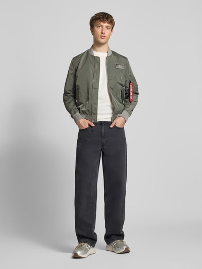 Männermodel trägt Regular Fit Bomberjacke mit Logo-Patch von Alpha Industries in Olivgrün, Stehkragen, Reißverschlusstaschen, Ganzkörperansicht von vorne.