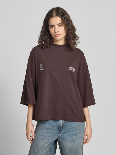 Kobiece model prezentuje Oversized T-Shirt mit Rundhalsausschnitt od Karo Kauer z bawełny, nadruk z logo, okrągły dekolt, krój oversized, długość tyłu w rozmiarze S: 59 cm, brąz i niebieski, widok całej sylwetki z przodu