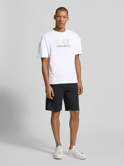Męski model nosi T-shirt mit Logo und Rundhalsausschnitt von EA7 Emporio Armani, bawełna, krój regular fit, logo, okrągły dekolt, długość tyłu w rozmiarze M: 68 cm, cała sylwetka z przodu.