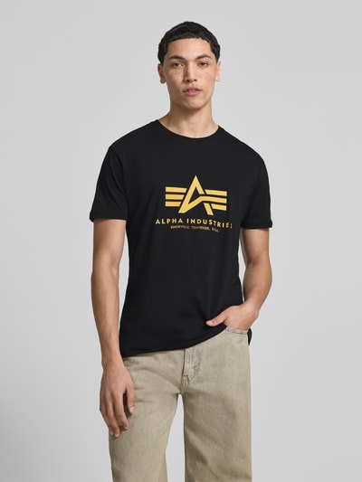 Männermodel trägt T-Shirt mit Label-Detail Modell 'Basic' von Alpha Industries aus Reine Baumwolle, Gerader-Schnitt, Rundhalsausschnitt, schwarze Farbe mit gelbem Aufdruck, Rückenlänge bei Größe M: 67 cm, Ganzkörperansicht von vorne.
