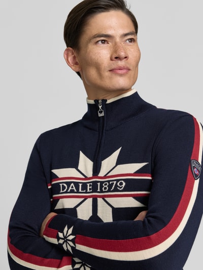 Männermodel trägt Wollpullover mit Label-Print Modell 'Nale' von Dale of Norway in Dunkelblau mit roten und weißen Streifen, Nahaufnahme Oberkörper, gerader Schnitt, Troyer-Kragen, reine Wolle, KI-modifiziertes Bild.