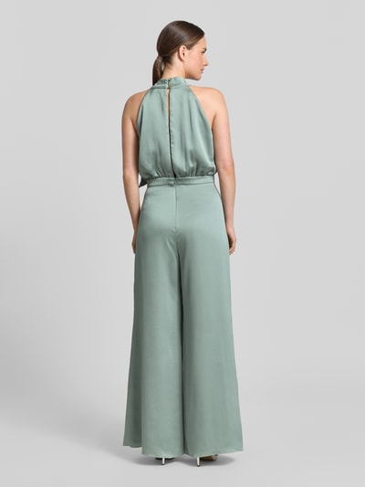 Kobiece model prezentuje Jumpsuit mit Schluppe und rückseitigem Knopfverschluss od Swing, satynowy materiał, krój slim fit, kolor miętowy, pasek w talii, długość całkowita 152 cm, długość nogawki wewnętrznej 82 cm, szerokość w rozmiarze 36: 66 cm, długość pleców 37 cm, zapięcie na guziki, haczyk i zamek z tyłu, widok całej sylwetki z tyłu.