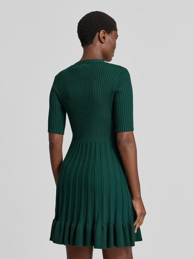 Frauenmodel trägt Strickkleid in Minilänge mit Viskose-Anteil von Christian Berg Woman in Dunkelgrün, Ripp-Optik, Rüschensaum, taillierte Passform, kurzärmelig, Ganzkörperansicht Rückansicht.