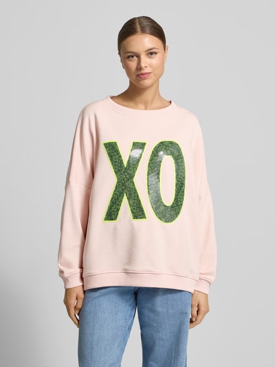 Frauenmodel trägt Oversized Sweatshirt mit Pailettenbesatz Modell 'XO' von miss goodlife in Rosa mit grünem Schriftzug, Baumwolle, Rundhalsausschnitt, elastische Abschlüsse, Ganzkörperansicht von vorne.