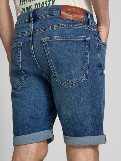 Männermodel trägt Slim Fit Jeansshorts aus Baumwoll-Mix Modell 'HAMPTON' von Calvin Klein Jeans in blauer Denim-Optik, 5-Pocket-Design, Label-Patch, Beinabschluss mit Umschlag, Ganzkörperansicht Rückansicht.