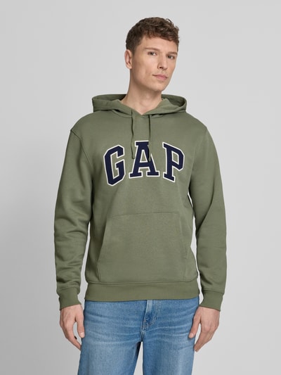 Männermodel trägt Hoodie mit Kängurutasche von GAP in Grün, Baumwoll-Mix, lockere Passform, Label-Stitching, gerippte Abschlüsse, Ganzkörperansicht von vorne.