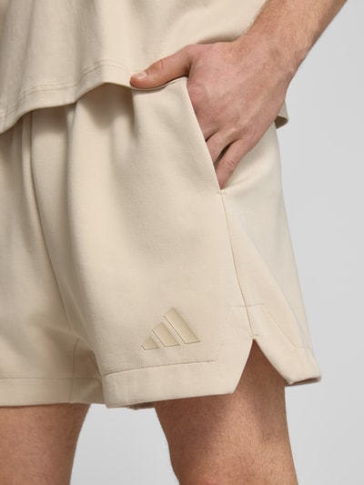 Mężczyzna nosi Regular Fit Sweatshorts mit Modal und Baumwoll-Anteil od adidas Sportswear, efekt: jednolity kolor, detal z logo, wpuszczane kieszenie, elastyczny ściągacz z tunelem, mieszanka materiałów, jasny beż, gładka tekstura, zbliżenie na dolną część nogi