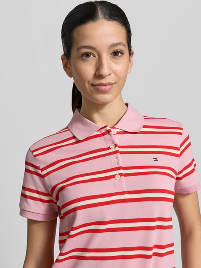 Frauenmodel trägt Regular Fit Poloshirt aus Baumwoll-Mix von Tommy Hilfiger, Optik: Unifarben, Muster: gestreift, Logo-Detail, Polokragen, kurze Knopfleiste, Kurzarm, Ärmelabschluss: gerippt, Rumpfabschluss: Tunnelzug, Materialverarbeitung: Piqué, Material: Baumwoll-Mix, Farben rosa und rot, Nahaufnahme Oberkörper.