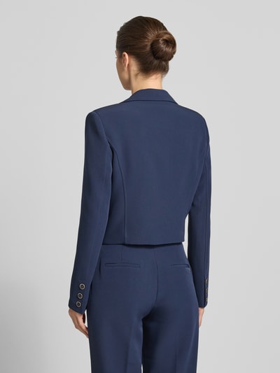 Frauenmodel trägt Cropped Blazer mit Reverskragen Modell 'Leila' von Guess in Dunkelblau, Elastischer-Material-Mix, Rückenansicht, Ganzkörperansicht Rückansicht, Pattentaschen, Knopfverschluss, Rückenlänge 47 cm, Brustweite 42 cm, Ärmellänge 64 cm.