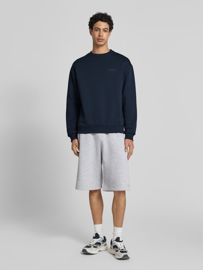 Męskimodel nosi Sweatshirt mit Rundhalsausschnitt und Logo od REVIEW, mieszanka bawełny, krój regular fit, okrągły dekolt, logo, długość tyłu w rozmiarze M: 70 cm, długość rękawów w rozmiarze M: 62 cm, ciemnogranatowy, gładka faktura. Ganzkörperansicht von vorne.