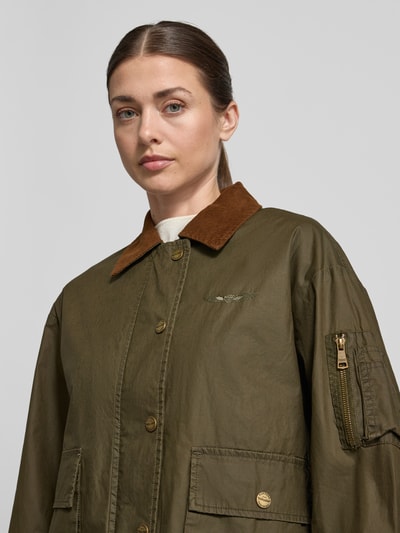 Frauenmodel trägt Jacke aus Baumwolle mit Pattentaschen von Original Bombers in Olive mit braunem Umlegekragen, Regular-Fit, Nahaufnahme Oberkörper. Rückenlänge 60 cm, Schulterbreite 52 cm, Brustweite 58 cm, Ärmellänge 57 cm, sichtbare Knopfleiste und Pattentaschen, strukturierte Baumwoll-Textur.