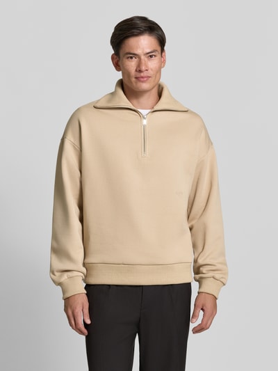 Męski model nosi Regular Fit Sweatshirt aus Baumwoll-Mix mit Troyer-Kragen od DRYKORN, mieszanka bawełny, krój regular fit, kołnierz zapinany, wyhaftowane logo, Długość tyłu w rozmiarze M: 64 cm, Długość rękawów w rozmiarze M: 57 cm, beżowa bluza o gładkiej fakturze, przód z zamkiem, Ganzkörperansicht von vorne