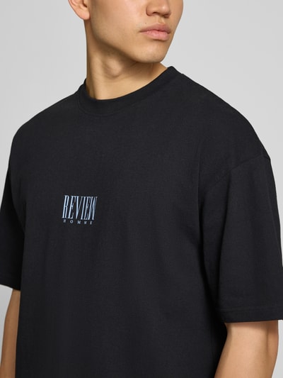 Männermodel trägt T-Shirt mit Logo-Print und Rundhalsausschnitt von REVIEW, schwarze Reine Baumwolle, Loose-Fit, Nahaufnahme Oberkörper.