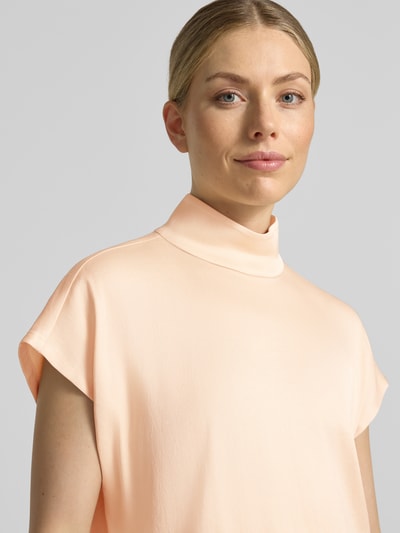 Frauenmodel trägt Blusenshirt mit Stehkragen Modell 'ALARIA' von DRYKORN in zartem Apricot, glatte Textur, Nahaufnahme Oberkörper, Material-Mix mit Stretch-Anteil, Regular Fit, Rückenlänge bei Größe 36: 60 cm.